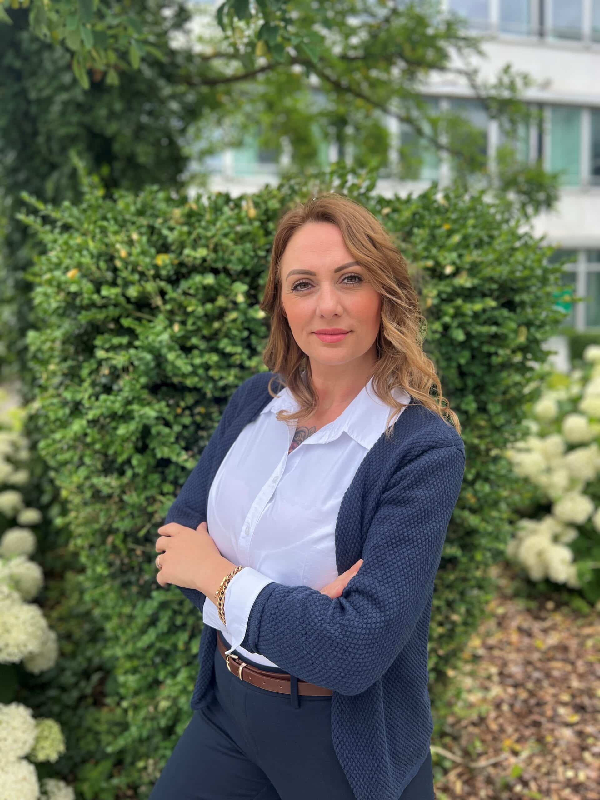 Eine Frau mit gewellten hellbraunen Haaren steht im Freien vor einer grünen Hecke. Gekleidet in eine weiße Bluse, einen marineblauen Blazer und eine Hose, strahlt sie Professionalität aus - und verkörpert damit perfekt den Geist von Lkm-Immobilien in München. Hinter ihr sind weiße Blumen und ein Gebäude zu sehen.