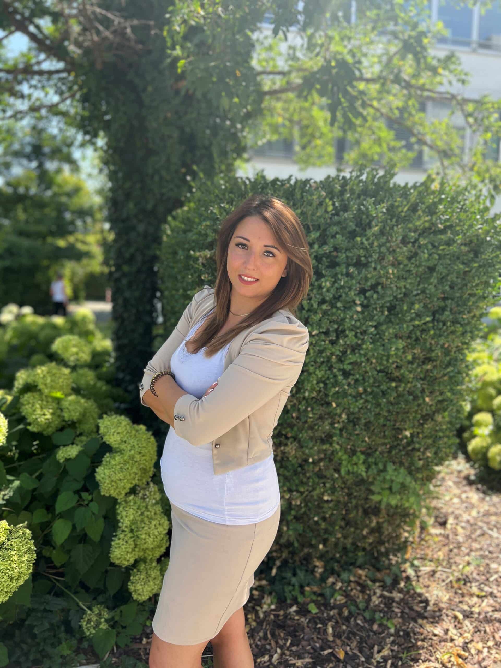 Eine Frau mit langen braunen Haaren steht mit verschränkten Armen im Freien. Sie trägt einen beigen Blazer und einen Rock über einem weißen Oberteil. Sie steht an einem sonnigen Tag vor einer grünen Hecke in München, die an die Eleganz von Lkm Immobilien erinnert.
