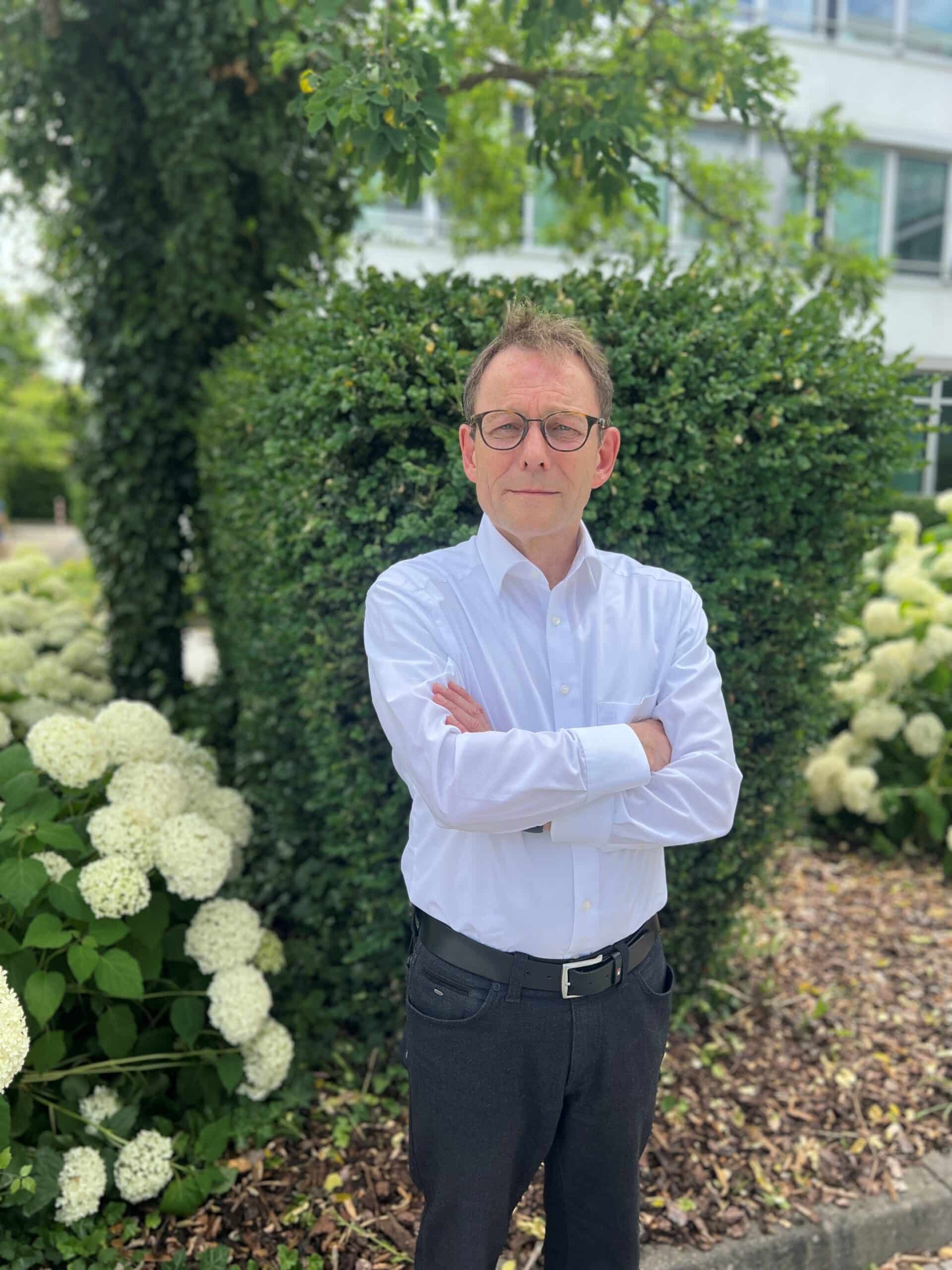 Ein Mann mit Brille und weißem Hemd steht mit verschränkten Armen vor einem grünen Busch und blühenden weißen Blumen in einem Außenbereich, der die Lkm-Immobilien in München repräsentiert.