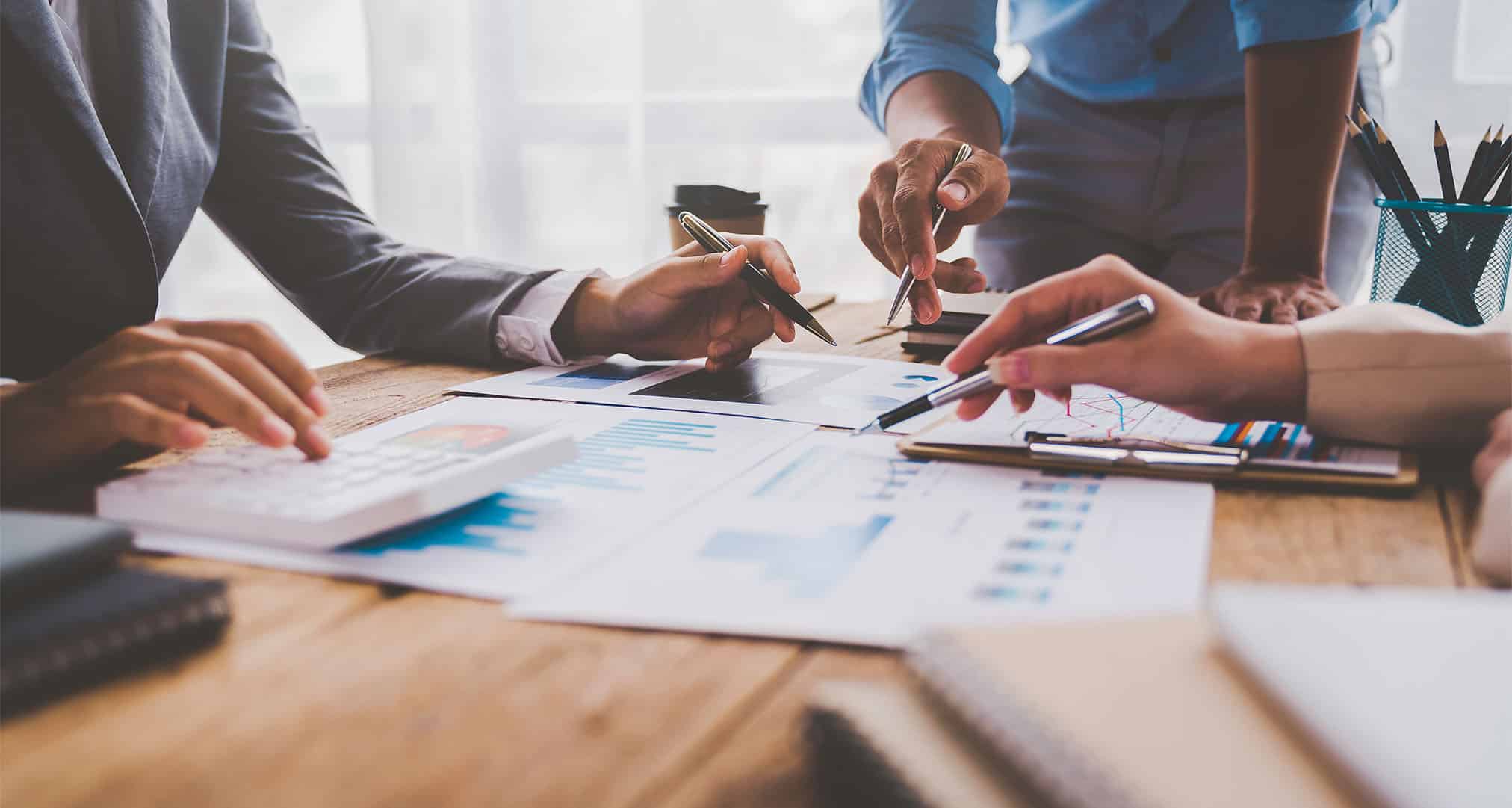 Drei Personen sitzen an einem Holztisch in München und analysieren die Diagramme und Tabellen von Immobilien auf Papier. Papiere, Stifte und ein Taschenrechner sind ausgebreitet, eine Person steht und zeigt auf ein Dokument, das den Fokus bei Lkm Immobilien widerspiegelt.